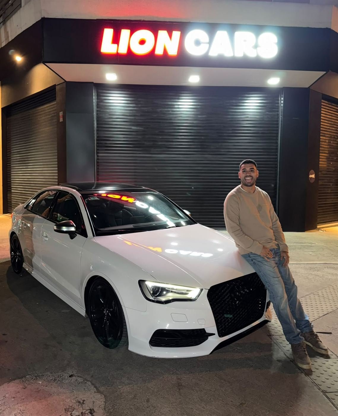 Nacho apoyado en un Audi blanco frente al cartel iluminado de Lion Cars
