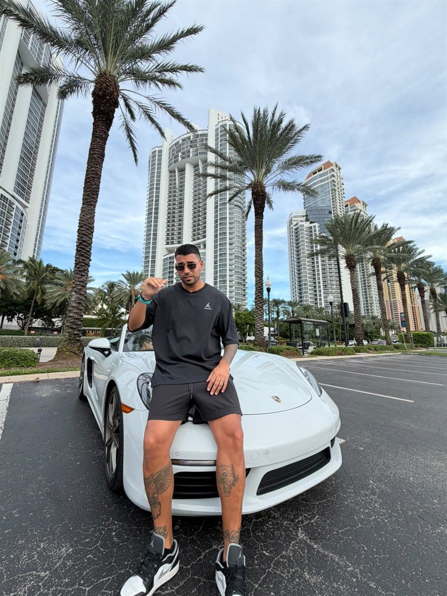 Nacho sentado sobre un Porsche blanco, con palmeras y edificios de fondo